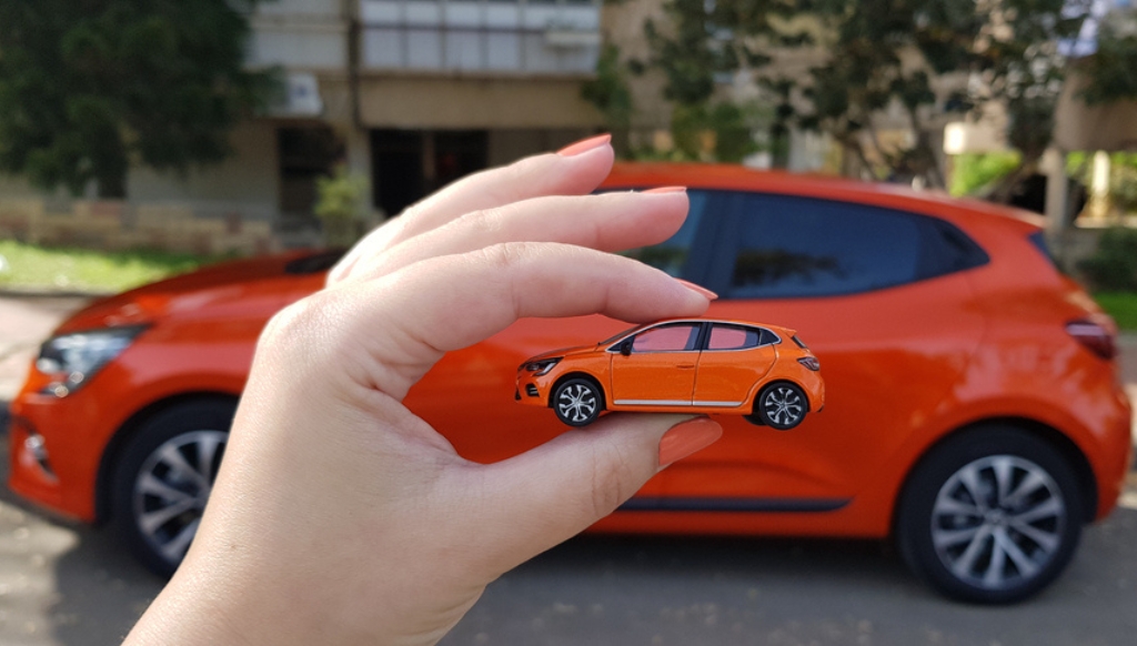 A female hand holds a miniature version of an orange salary sacrifice car in front of the real life version.